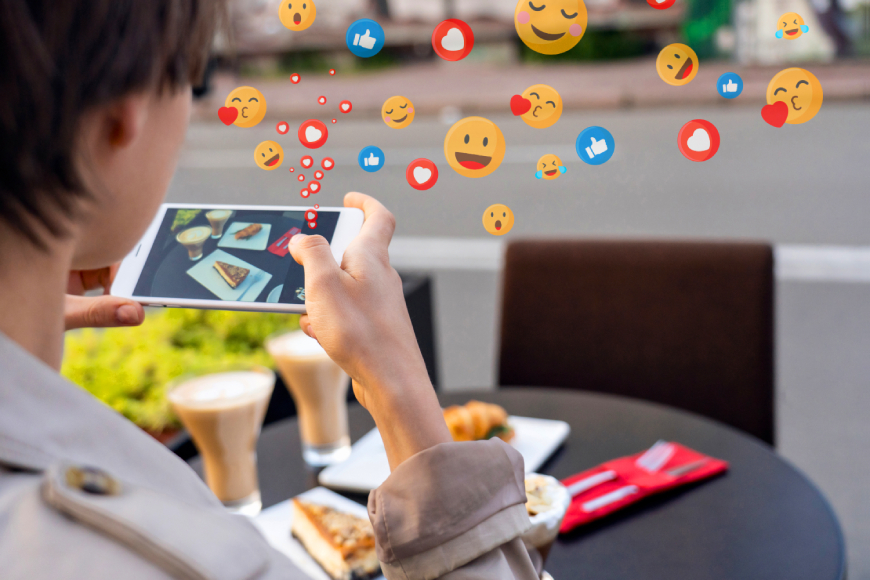 Girl holding smartphone taking photo of food on outdoor restaurant table with social media thumbs up, emoji reactions and hearts