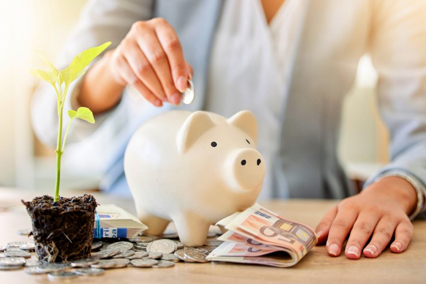 Hand putting coins into piggy bank on desk with money notes and growing plant