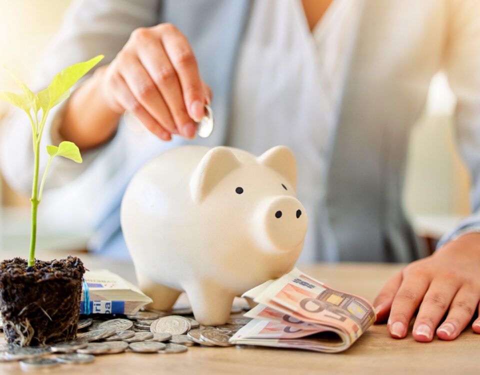 Hand putting coins into piggy bank on desk with money notes and growing plant