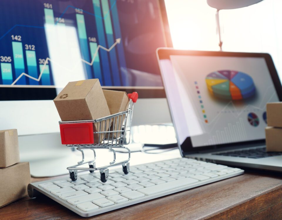 Miniature shopping cart full of cardboard boxes on computer keyboard beside laptop