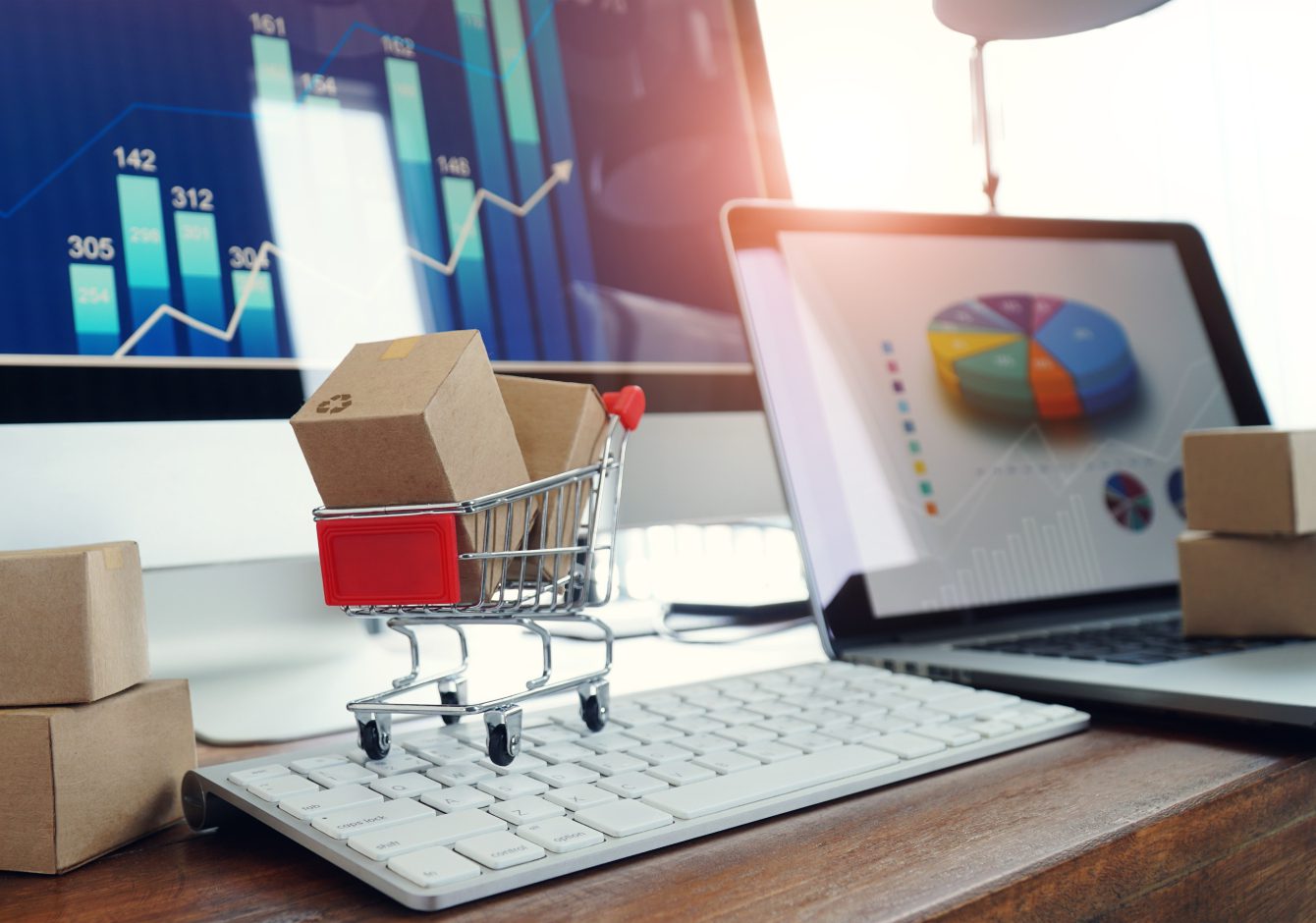 Miniature shopping cart full of cardboard boxes on computer keyboard beside laptop