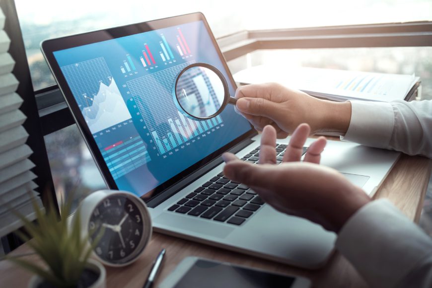 Hand holding magnifying glass in front of laptop screen with graphs on it