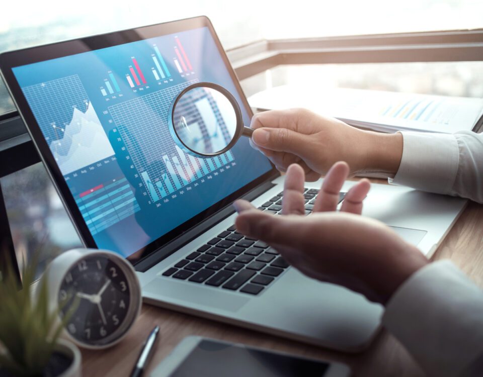 Hand holding magnifying glass in front of laptop screen with graphs on it