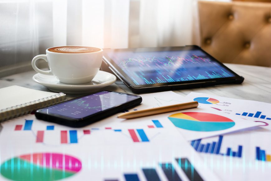 iPad tablet on desk beside coffee cup and smartphone with charts and graphs on paper