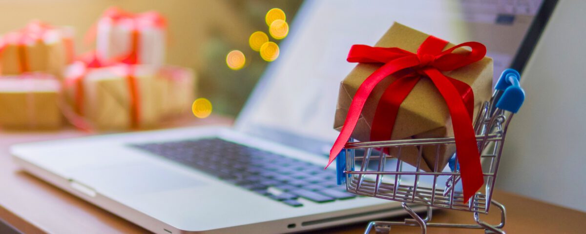 Mini shopping cart with gift inside it on table beside laptop with Christmas tree in background