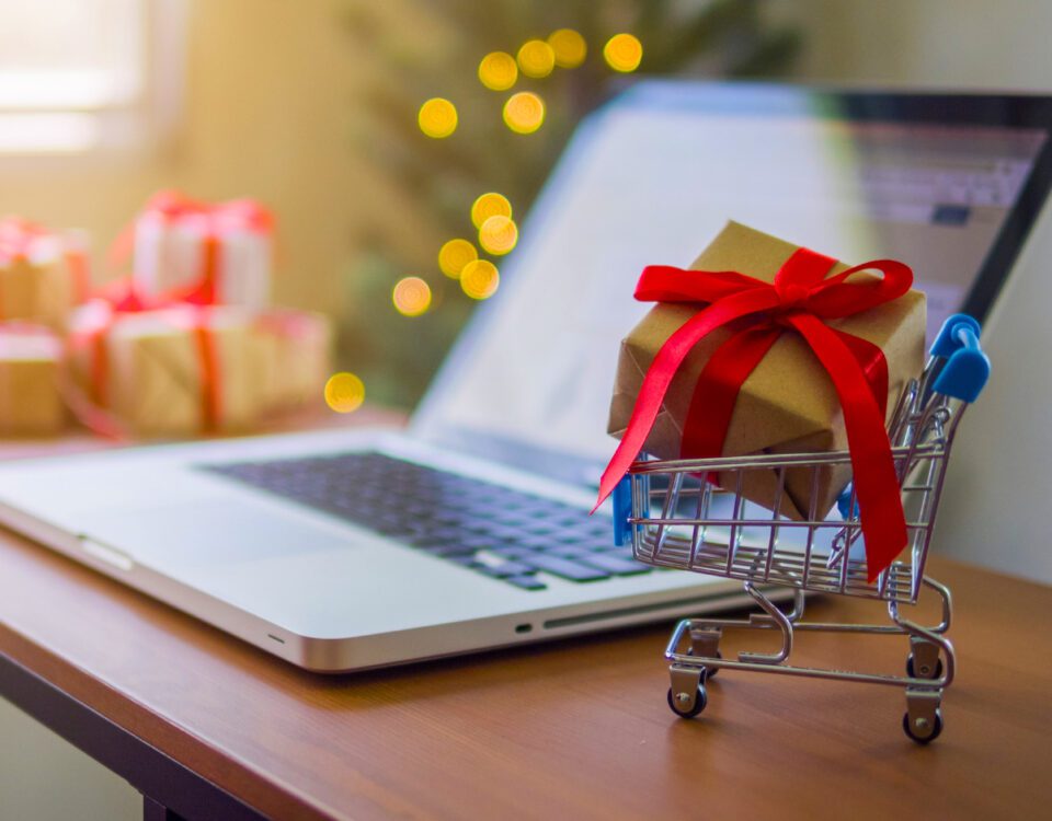 Mini shopping cart with gift inside it on table beside laptop with Christmas tree in background