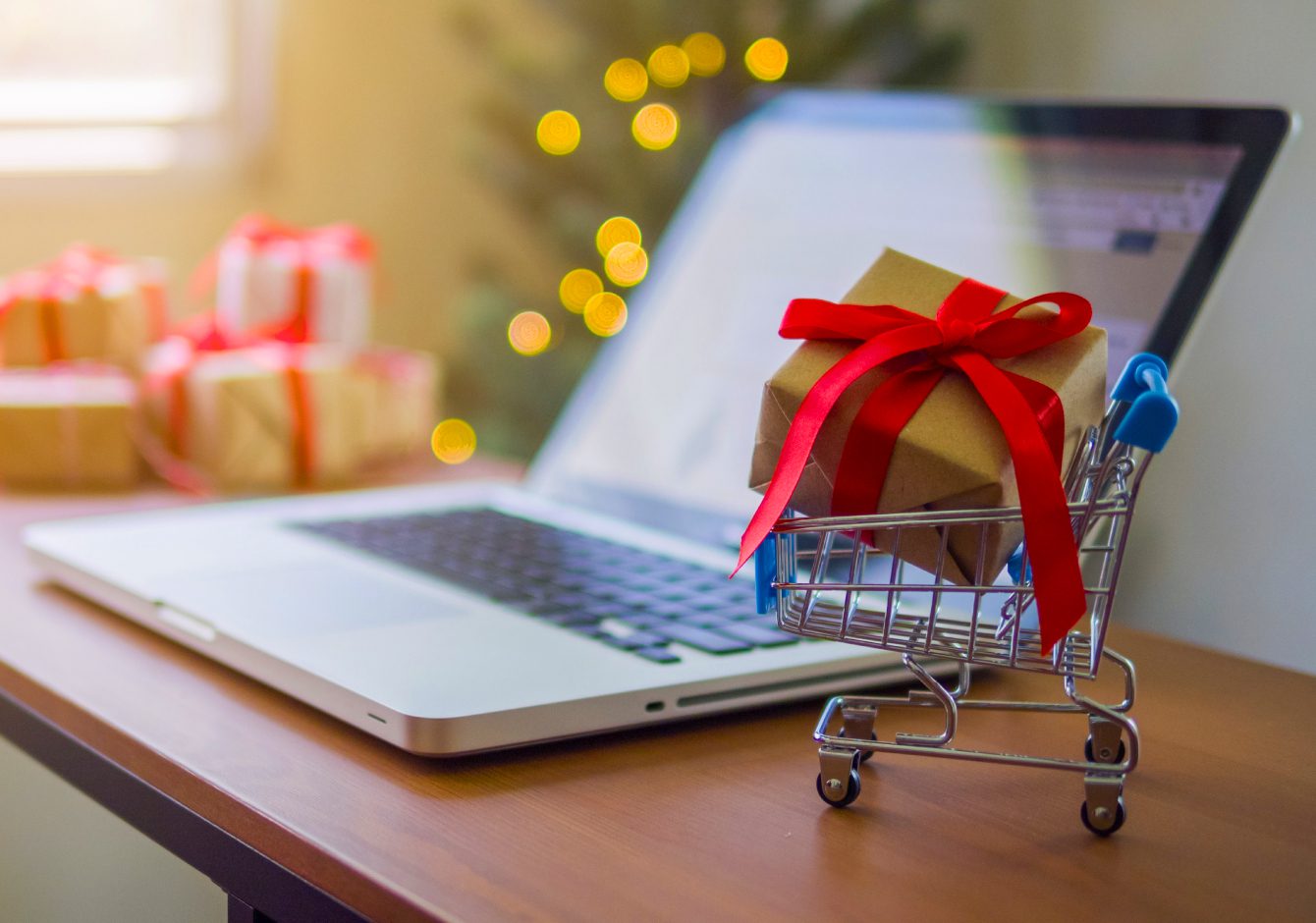 Mini shopping cart with gift inside it on table beside laptop with Christmas tree in background
