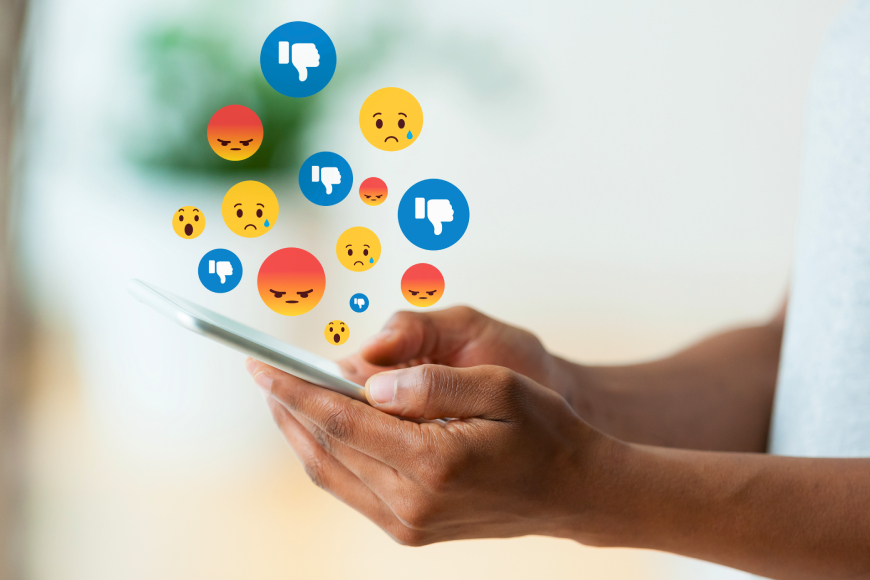 Hands holding smartphone with thumbs down, unhappy and angry emojis coming out of screen