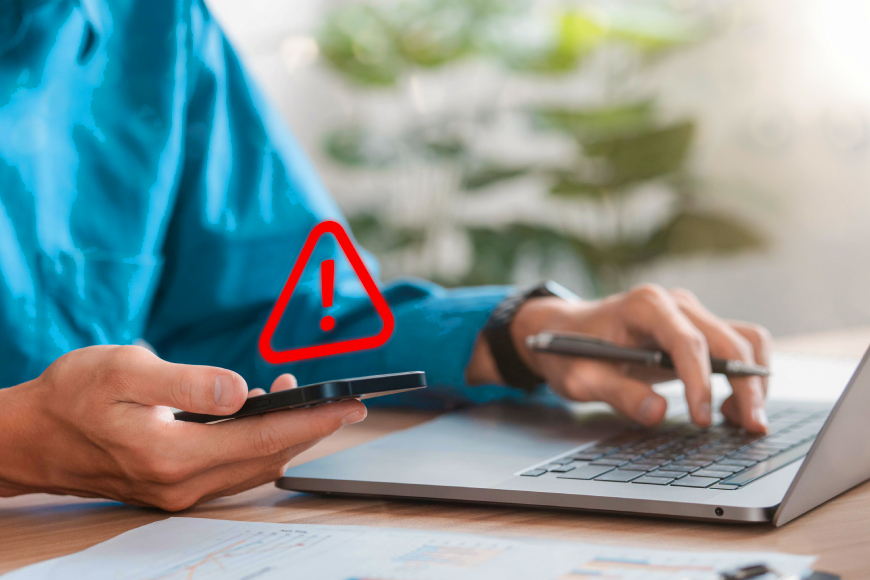 Hands using smartphone and laptop at desk with red exclamation mark alert symbol above phone