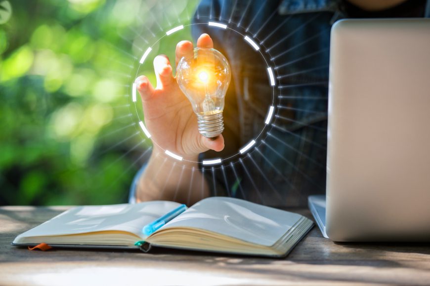 Person sitting at desk with book and laptop holding a glowing lightbulb in their hand