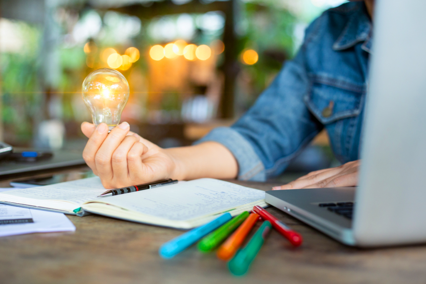 Person sitting at desk with notepad and laptop holding a glowing lightbulb in their hand