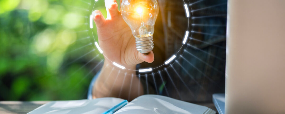 Person sitting at desk with book and laptop holding a glowing lightbulb in their hand