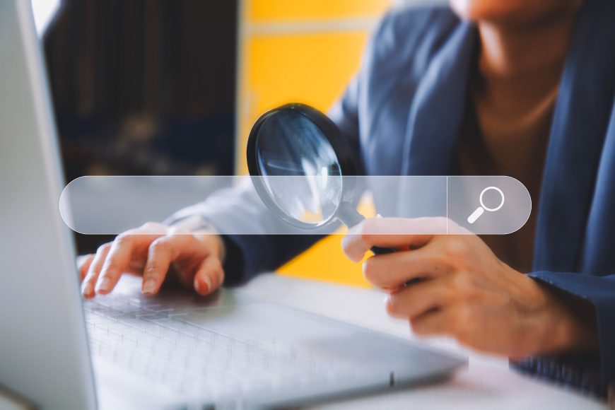 Person sitting at desk holding magnifying glass to laptop screen with search bar