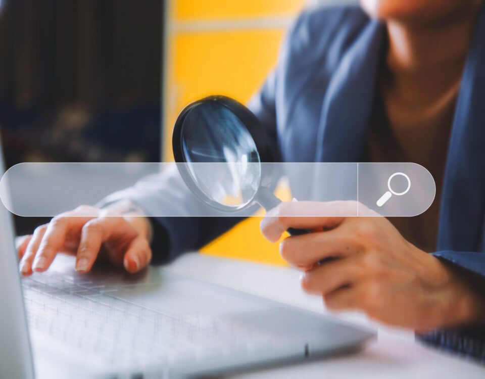 Person sitting at desk holding magnifying glass to laptop screen with search bar