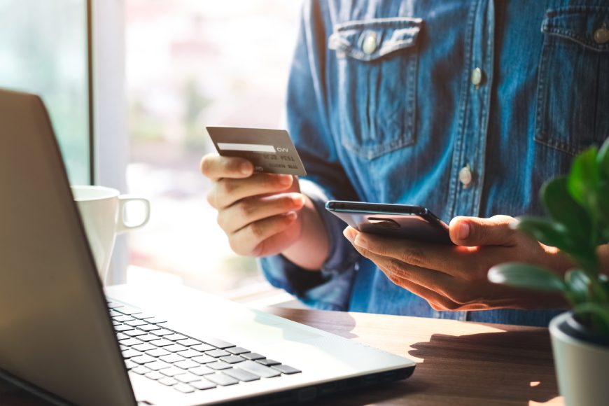 Person holding credit card and using smartphone in front of laptop