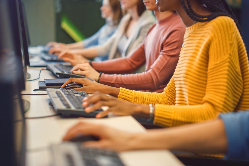 Row of people sitting at desk in office typing on computer keyboards