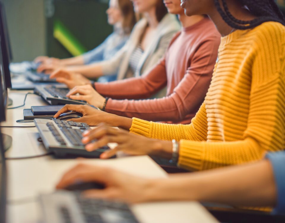 Row of people sitting at desk in office typing on computer keyboards