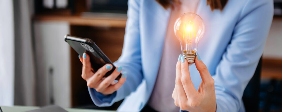 Woman sitting at desk with laptop holding smartphone and glowing lightbulb