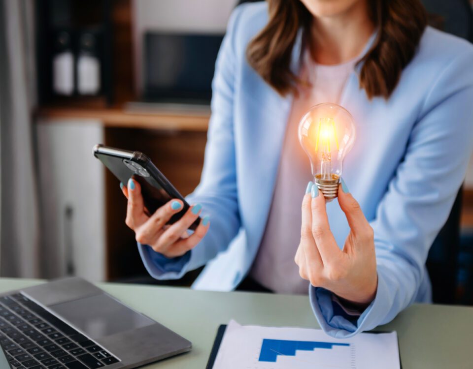 Woman sitting at desk with laptop holding smartphone and glowing lightbulb