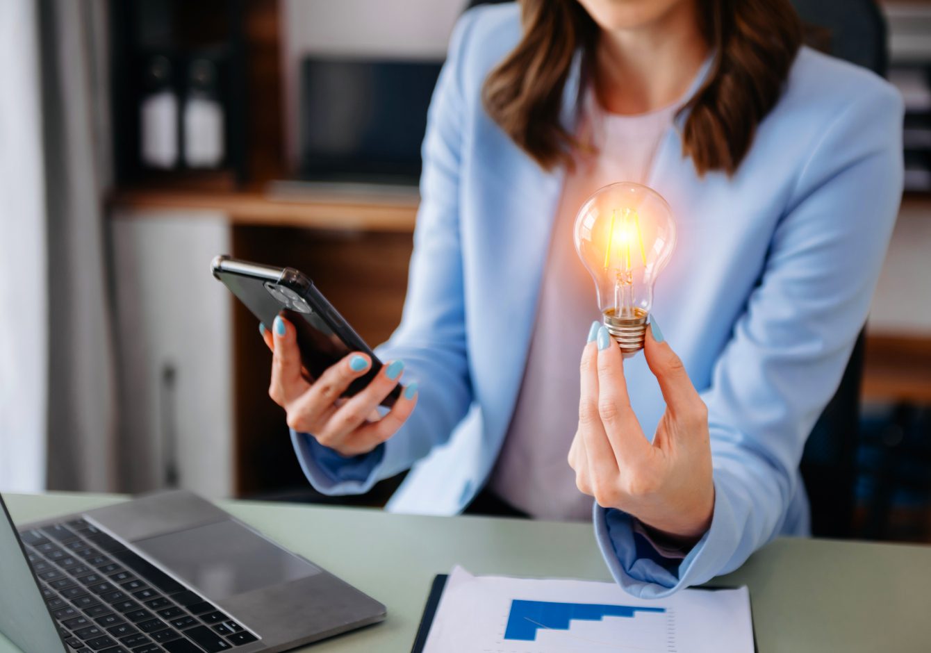 Woman sitting at desk with laptop holding smartphone and glowing lightbulb