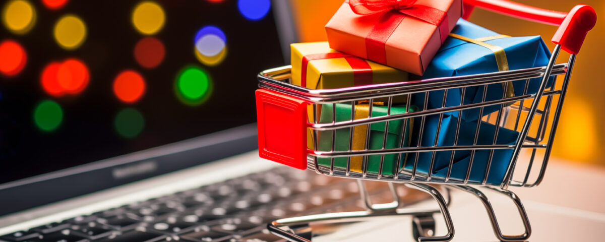 Miniature shopping cart filled with Christmas gifts sitting on laptop keyboard with festive lights behind