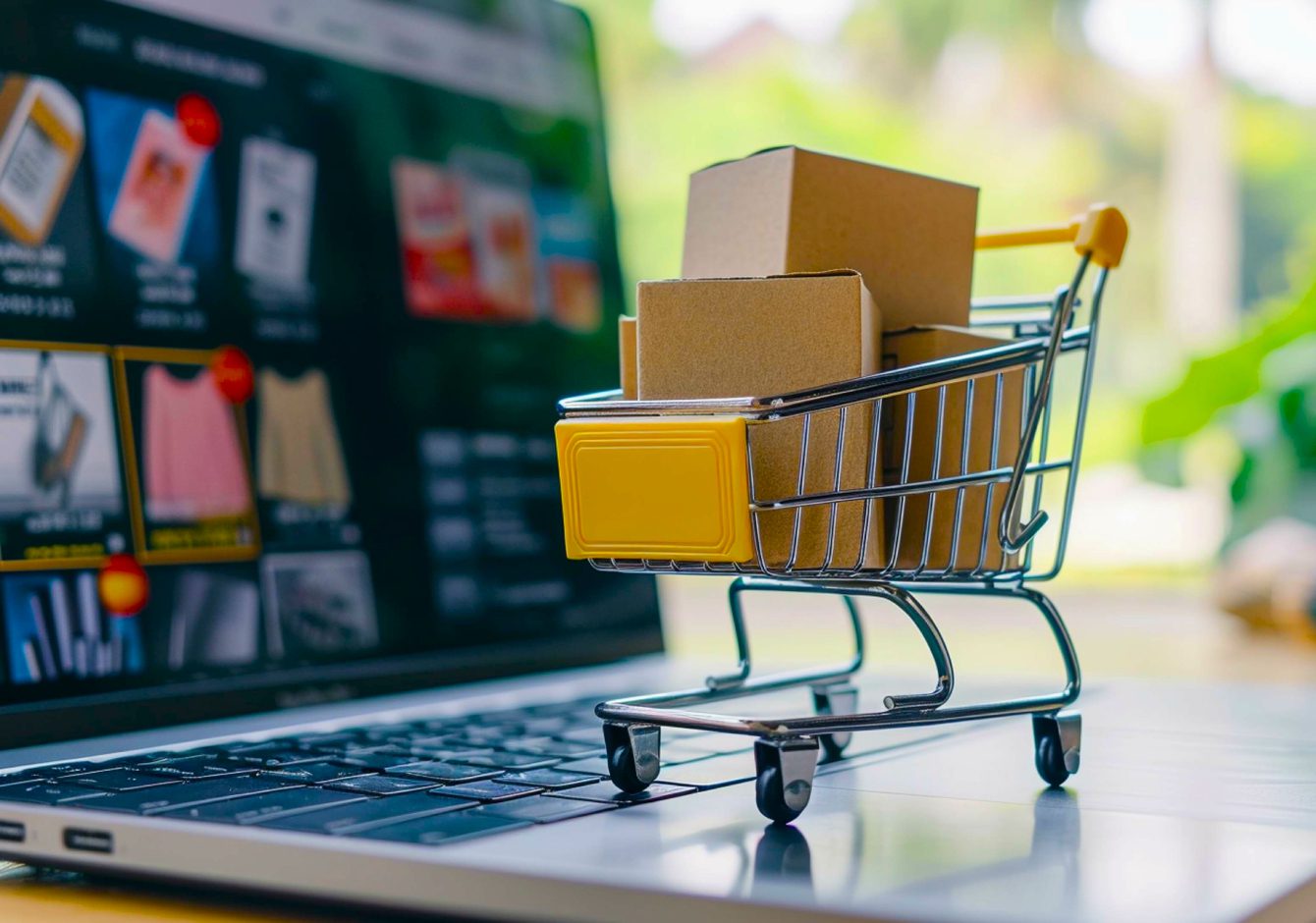 Miniature shopping cart full of boxes sitting on laptop with eCommerce store on screen