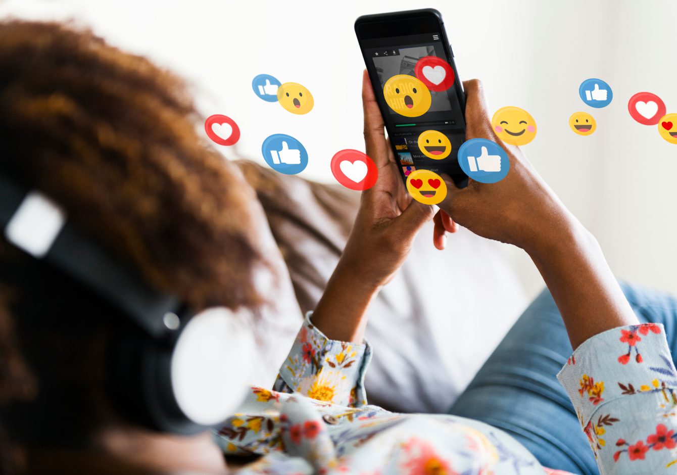Woman lounging on sofa using smartphone with social media reaction emojis around