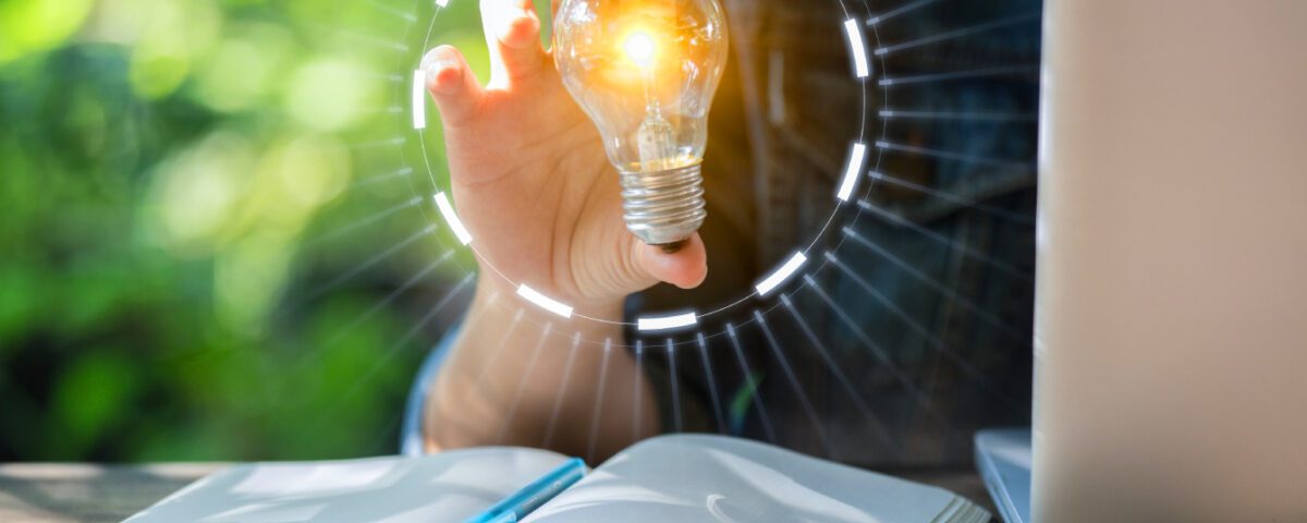 Person sitting at desk with laptop and notebook holding a glowing lightbulb in one hand