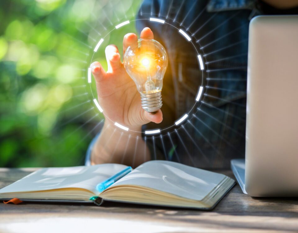 Person sitting at desk with laptop and notebook holding a glowing lightbulb in one hand