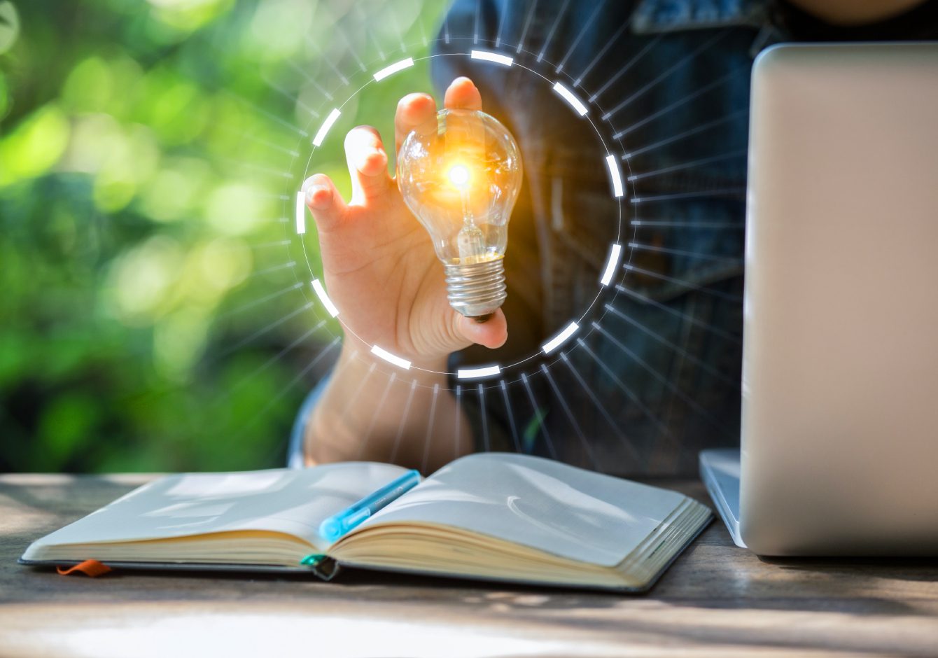 Person sitting at desk with laptop and notebook holding a glowing lightbulb in one hand