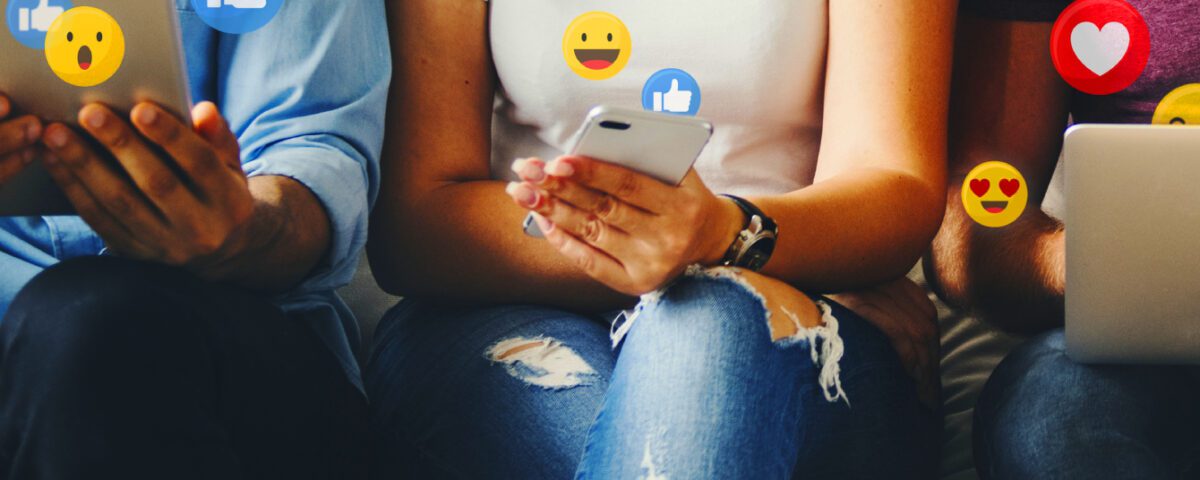 Three people sitting on a sofa using a tablet, smartphone and laptop with social media reaction emojis around