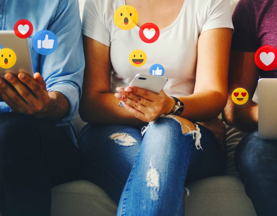 Three people sitting on a sofa using a tablet, smartphone and laptop with social media reaction emojis around