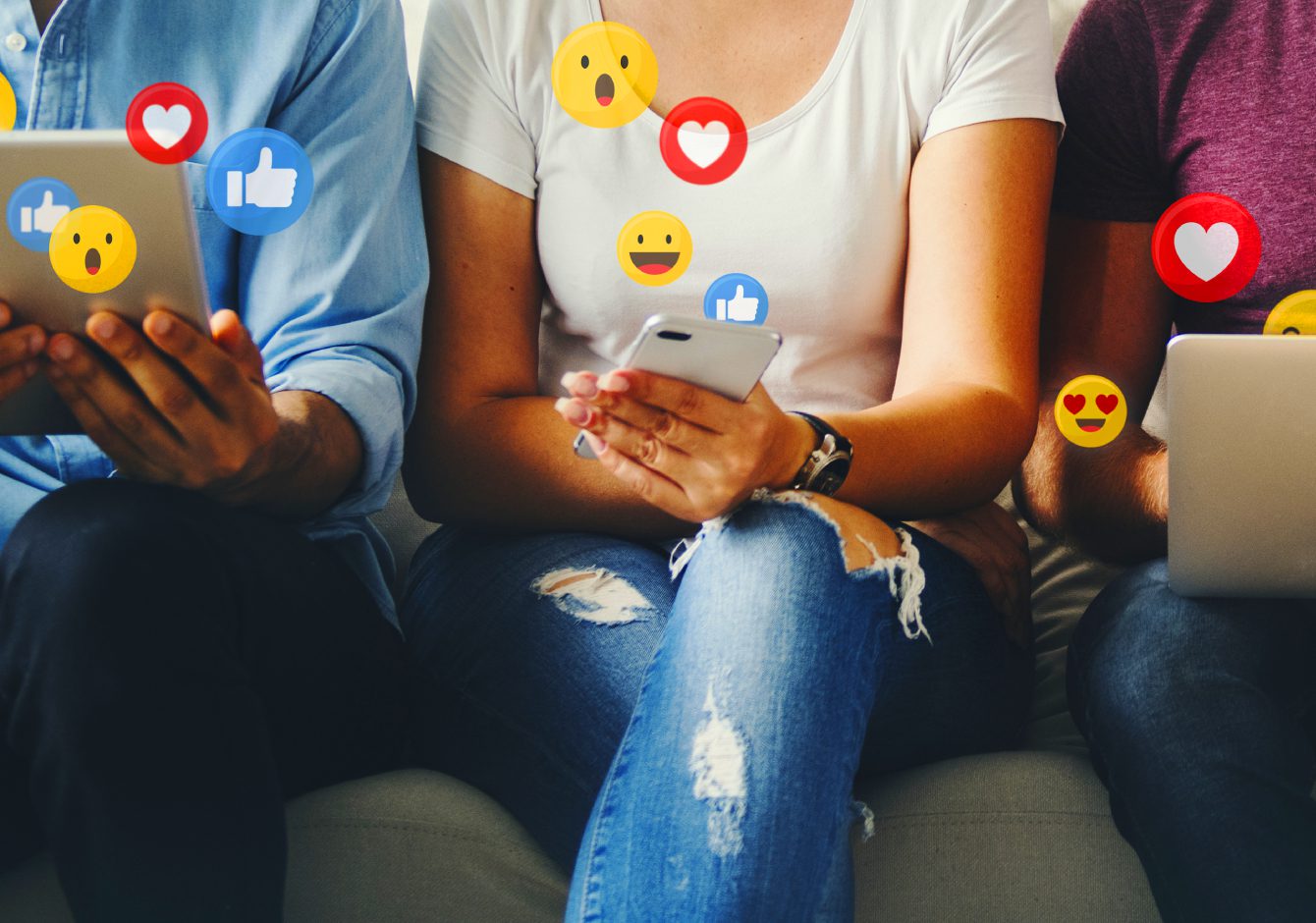 Three people sitting on a sofa using a tablet, smartphone and laptop with social media reaction emojis around