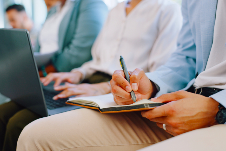 Row of people sitting in office typing on a laptop and writing in a notebook