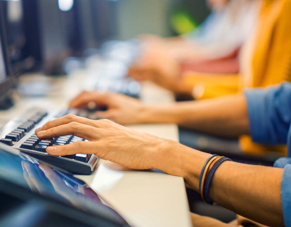 Row of people sitting at desk in office typing on computer keyboards