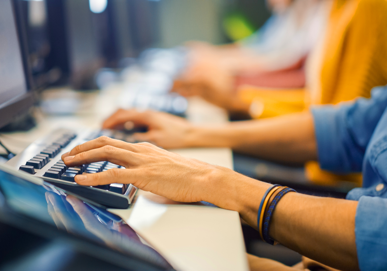 Row of people sitting at desk in office typing on computer keyboards