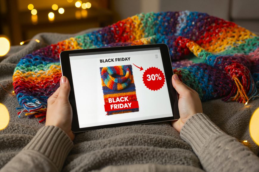 Person sitting under comfy rainbow blanket using iPad tablet with Black Friday Sale on screen and glowing lights behind