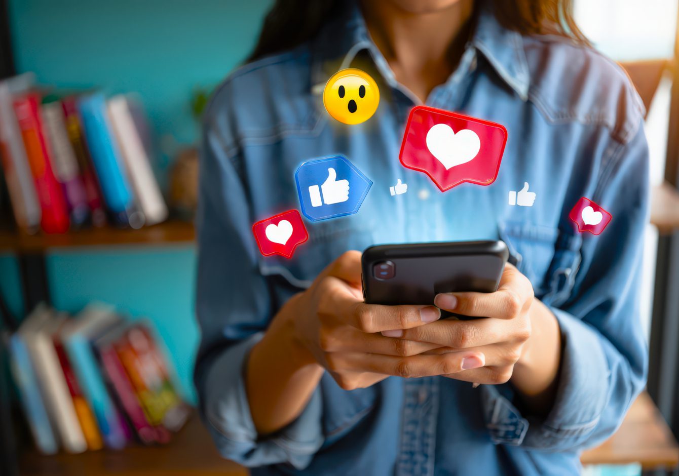 Woman in blue shirt holding smartphone with social media reaction emojis around